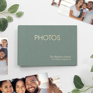 May include: A sage green photo album with the word "PHOTOS" in gold lettering. The album also has the text "The Stephens Family" and "Our Summer Adventures" in gold. Family photos are in the background.