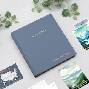May include: A blue hardcover journal with the text "NATIONAL PARKS" and "Thompson Family Adventures" on the cover. The journal is surrounded by postcards with images of national parks.