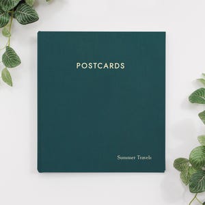 May include: A dark teal hardcover book with the word "POSTCARDS" in gold lettering at the top and "Summer Travels" at the bottom. The book is surrounded by green leafy plants.