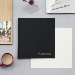 May include: A black hardcover journal with a gold inscription that reads "Your Custom Text Here Your Custom Text Here". The journal is on a light gray surface with a white sheet of paper next to it.