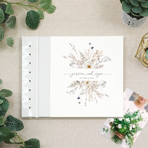 May include: A white wedding guest book with a white ribbon closure. The cover features a watercolor illustration of dried flowers and the text "Jessica and Ryan, October 26, 2024".