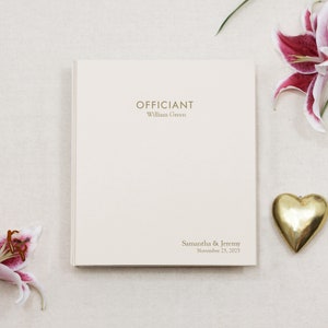 May include: A white card with the text "Officiant" and "William Green" printed in gold. Below the text is the text "Samantha & Jeremy" and "November 23, 2025" printed in gold.