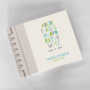May include: A personalized school years photo album with a cream-colored cover. The cover features a colorful alphabet design in green, blue, and gray, with the name "FRANCO MARCUS" and "SCHOOL YEARS" printed below. The album is spiral-bound.