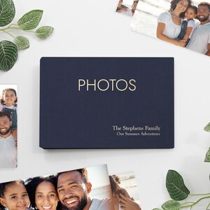 May include: A navy blue photo album with the word "PHOTOS" in gold lettering. Below, the text reads "The Stephens Family" and "Our Summer Adventures". The album is rectangular and surrounded by family photos and greenery.