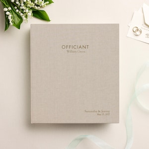 May include: A square, light beige linen-covered book with gold foil text on the front cover. The text reads "OFFICIANT William Green" at the top and "Samantha & Jeremy May 15, 2027" at the bottom right corner. The cover has a visible fabric texture.