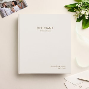 May include: A white hardcover book with gold foil text that reads "OFFICIANT William Green" at the top and "Samantha & Jeremy May 15, 2027" at the bottom right corner. The book is positioned next to a small photograph and white flowers.