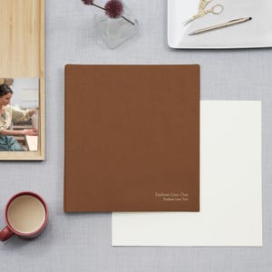 May include: A square, brown, leather-like binder featuring the text "Emboss Line One" and "Emboss Line Two" printed in the bottom right corner. The binder is positioned on a light gray surface next to a white sheet of paper.