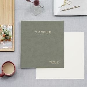 May include: A green, textured, hardcover journal with the text "YOUR TEXT HERE" on the front cover. The journal is lying on a light gray surface with a white sheet of paper underneath it.