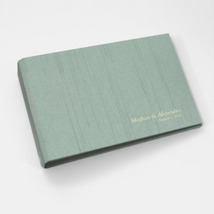 May include: A sage green wedding guest book with the text "Meghan & Alejandro August 1, 2022" printed in gold.