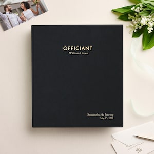 May include: A black hardcover book with gold foil text that reads "OFFICIANT", "William Green", "Samantha & Jeremy", and "May 15, 2027". The book is positioned centrally, accompanied by a small photograph of a couple and white flowers.