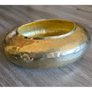 May include: A hammered gold-colored bowl with a wide, oval shape. The interior of the bowl is a bright gold, contrasting with the textured exterior. The bowl is likely made of metal and has a decorative, handcrafted appearance.