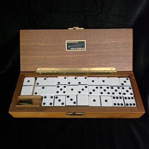 May include: A wooden domino set in a rectangular box with a brass hinge and latch. The interior contains white dominoes with black pips and a small compartment for metal pegs. A black label on the lid reads Handcrafted Five-Up Company.