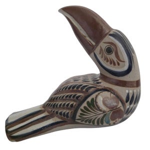 May include: A ceramic toucan figurine featuring a beige base color with hand-painted details in brown, black, and green. The bird has a large, curved brown beak, stylized feather patterns on its wings, and striped markings on its tail feathers.