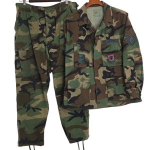 May include: A camouflage uniform set consisting of a long-sleeved button-up jacket and matching trousers. The jacket features U.S. AIR FORCE text, two chest pockets, and embroidered patches on the chest and sleeve. The pattern is woodland camo.