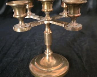 May include: A polished brass candelabra featuring a central stem rising from a circular base, supporting four arms that extend outward to hold individual candles. The metal surface has a warm, metallic gold tone with a smooth, reflective finish.