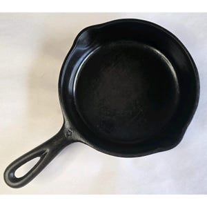 May include: A black cast iron frying pan with a single handle. The pan has a round cooking surface and a small pouring spout. The handle has a hole for hanging. The pan is made of heavy-duty cast iron.