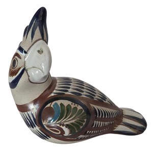 May include: A ceramic bird figurine featuring a cream-colored base with hand-painted details in dark blue, brown, and green. The bird has a prominent crest, a curved beak, and intricate feather patterns covering its body, wings, and tail feathers.