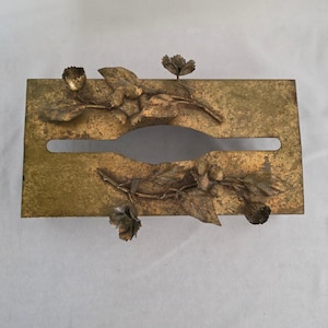 May include: A rectangular gold-toned metal tissue box cover featuring a weathered, distressed finish. The top surface is decorated with two raised, three-dimensional metal floral branches with leaves and small buds extending across the center.