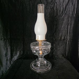 May include: A clear glass oil lamp with a white frosted glass shade and a brass-colored metal frame. The lamp has a round base and a decorative, patterned glass body. The lamp is designed to hold oil and provide light.