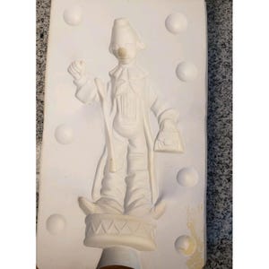 Könnte beinhalten: Eine weiße Keramik-Clown-Figur-Form. Der Clown trägt einen Hut, einen Mantel und hält eine Tasche. Die Form hat eine strukturierte Oberfläche mit mehreren runden Vertiefungen.