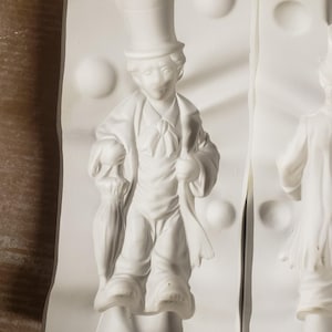 May include: A white silicone mold featuring a relief design of a figure wearing a top hat and a coat, holding a closed umbrella. The mold has a smooth, matte surface with circular indentations and a clear, detailed impression of the character.