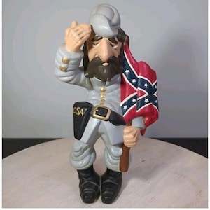 Puede incluir: Figura de un soldado de la Guerra Civil con uniforme gris, saludando con la mano derecha. Sostiene una bandera confederada con fondo rojo y estrellas azules. La figura tiene una larga barba y las letras "CSA" en su cinturón.