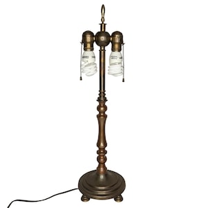 Può includere: Una lampada da tavolo color bronzo con una colonna decorativa tornita e una base rotonda. La lampada ha tre portalampada con catenelle e lampadine a risparmio energetico. La lampada è collegata a un cavo elettrico nero.