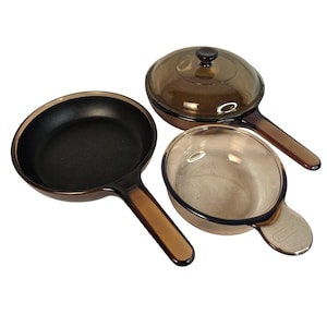 May include: A set of three Vision cookware pieces including a black-interior skillet with a long handle, a translucent amber glass pan with a matching lid, and a small amber glass bowl with a handle, each featuring the Vision brand logo.