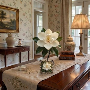 May include: A dining room scene featuring a large white magnolia flower arrangement in a decorative vase. The table is adorned with a floral runner, a lamp, books, and a small framed picture of a magnolia. The room has floral wallpaper and a painting.
