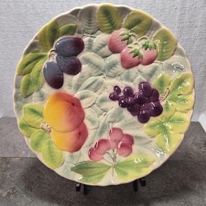May include: A decorative ceramic plate featuring a relief design of various fruits. The plate showcases plums, strawberries, grapes, cherries, and peaches, with green leaves. The plate is supported by a black stand.