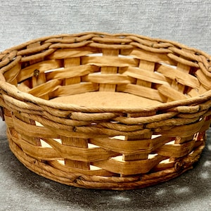 May include: A round, woven basket with a natural brown color. The basket has a sturdy construction with a woven pattern and a flat base. The rim is reinforced with a darker brown material. The basket is empty and sits on a gray surface.