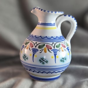May include: A white ceramic pitcher with a handle and spout, adorned with blue, yellow, and colorful floral patterns. The pitcher features a wavy blue line design and is likely used for serving beverages.