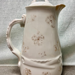 May include: A white ceramic pitcher with a lid, handle, and spout. The pitcher is decorated with gold-colored floral patterns. The handle and spout have ornate detailing. The pitcher is approximately 10 inches tall.