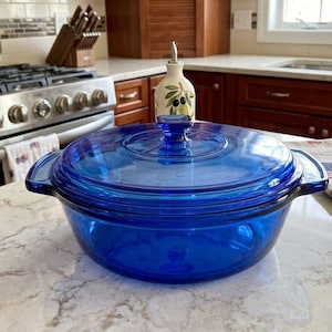 May include: A round, deep cobalt blue glass casserole dish with a matching glass lid. The dish features two integrated side handles and a central knob on the lid. The glass is translucent, allowing light to pass through the vibrant blue material.