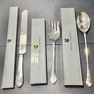 May include: A silver-plated knife, fork, and spoon set from Royal Limited Silver. Each piece of cutlery features a decorative shell-like motif at the base of the handle and is accompanied by its own individual light gray storage box with branding.