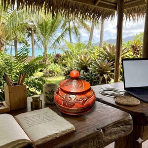 May include: An orange ceramic jar with a lid, featuring carved floral designs. The jar sits on a wooden table, alongside a notebook, pens, and a laptop. The background includes lush greenery and a view of the ocean.