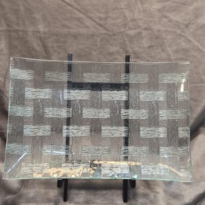 May include: Clear glass rectangular dish with a woven texture pattern. The dish is displayed on a black metal stand, showcasing its design. The glass has a slight curve on the edges.