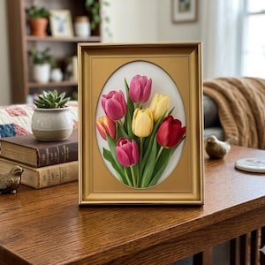 May include: A framed floral artwork featuring a bouquet of tulips in shades of red, pink, and yellow. The oval-shaped image is set against a white background and framed in a gold-colored rectangular frame. The artwork is displayed on a wooden table.