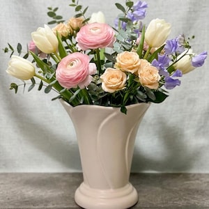 May include: A floral arrangement in a light pink vase. The bouquet includes white tulips, pink ranunculus, peach roses, and purple sweet peas, with green foliage. The vase has a curved design and a wide base.