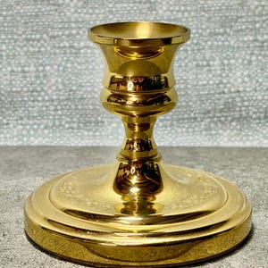 May include: A gold-colored candlestick holder with a wide, circular base and a flared top. The candlestick has a smooth, reflective surface and a classic design. The holder is made of metal and is suitable for a standard-sized candle.