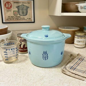 May include: A light blue ceramic Dutch oven with a lid, adorned with blue floral designs. A Pyrex measuring cup and a jar of baking powder are in the foreground. A vintage supermarket advertisement is in the background.