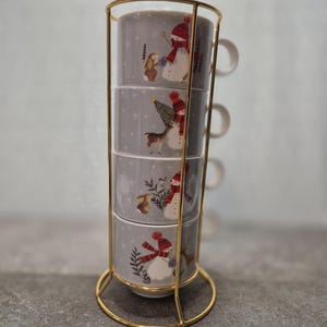 May include: A set of four gray ceramic mugs stacked in a gold-colored metal stand. Each mug features a winter scene with a snowman, deer, and forest elements. The mugs are designed with a handle and are suitable for hot beverages.