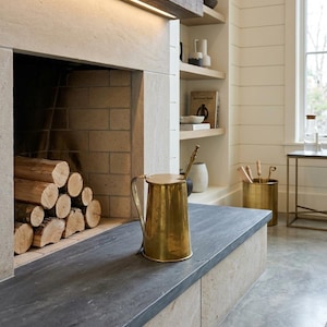 May include: A brass pitcher with a lid sits on a dark gray stone hearth in front of a fireplace. The fireplace has a light-colored stone surround and is filled with stacked firewood. A wooden beam with a light strip is above the fireplace.