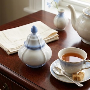 May include: A white and blue ceramic sugar bowl with a lid, a teacup and saucer, a teapot, and a small vase with blue flowers. The teacup contains tea, and there are two sugar cubes on the saucer. The items are on a dark wooden table.