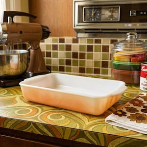 May include: A rectangular baking dish made of white milk glass with a gradient orange exterior finish. The dish features integrated handles on both short ends and a smooth, glossy surface. It is positioned on a kitchen counter near a mixer.
