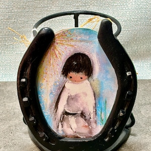 May include: A black horseshoe-shaped metal stand holding a circular painting. The painting depicts a figure in white clothing with black hair, set against a colorful background. Dried straw is attached to the top of the stand.