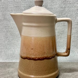May include: A ceramic pitcher with a lid, featuring a cream-colored top, transitioning to a light brown middle, and a darker brown base. The pitcher has a handle and a spout for pouring. The overall design is simple and functional.