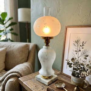 May include: A vintage-style table lamp with a frosted glass shade and a white base. The shade is adorned with delicate floral patterns. The lamp sits on a marble base with brass accents, creating a warm, inviting glow. The lamp is approximately 15 inches tall.