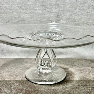 May include: A clear glass cake stand with a scalloped edge. The stand has a round base and a decorative stem. The cake stand is made of clear glass and is suitable for displaying cakes or other desserts.
