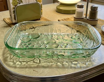 May include: A rectangular, light green glass baking dish with a fruit and leaf pattern. The dish has rounded corners and a beaded edge. It sits on a light-colored countertop, suggesting a vintage kitchen aesthetic.